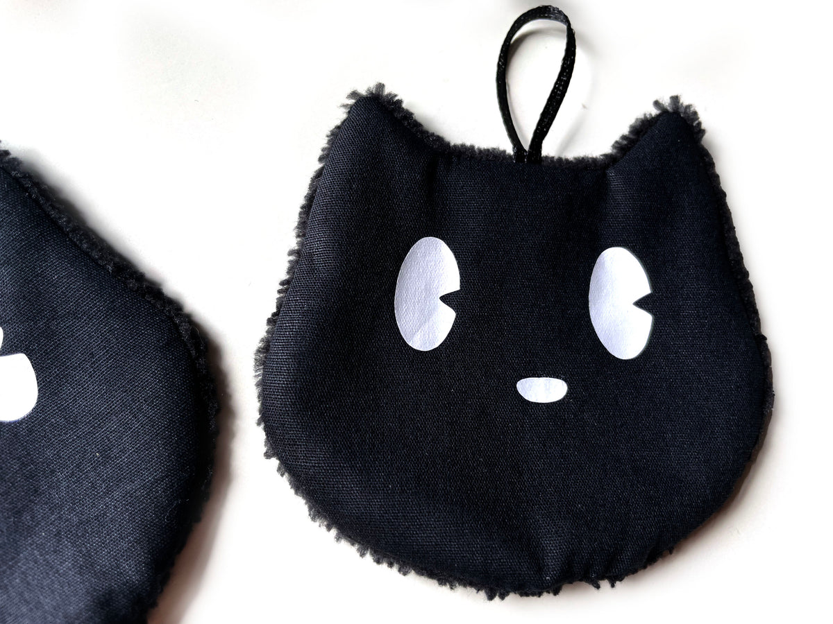 Detailansicht eines schwarzen waschbaren Abschminkpads in Katzenform mit weißen Augen, Satinschlaufe und flauschigem Rand