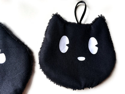 Detailansicht eines schwarzen waschbaren Abschminkpads in Katzenform mit weißen Augen, Satinschlaufe und flauschigem Rand