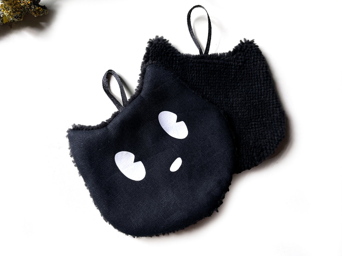 Nahaufnahme zweier schwarzer Abschminkpads in Katzenform: vorne Baumwolle mit weißem Gesicht, dahinter flauschiger Bambusfrottee und Satinschlaufen